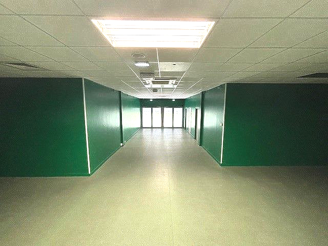 Long couloir vide aux murs verts et aux plafonds lumineux, menant à des portes vitrées.