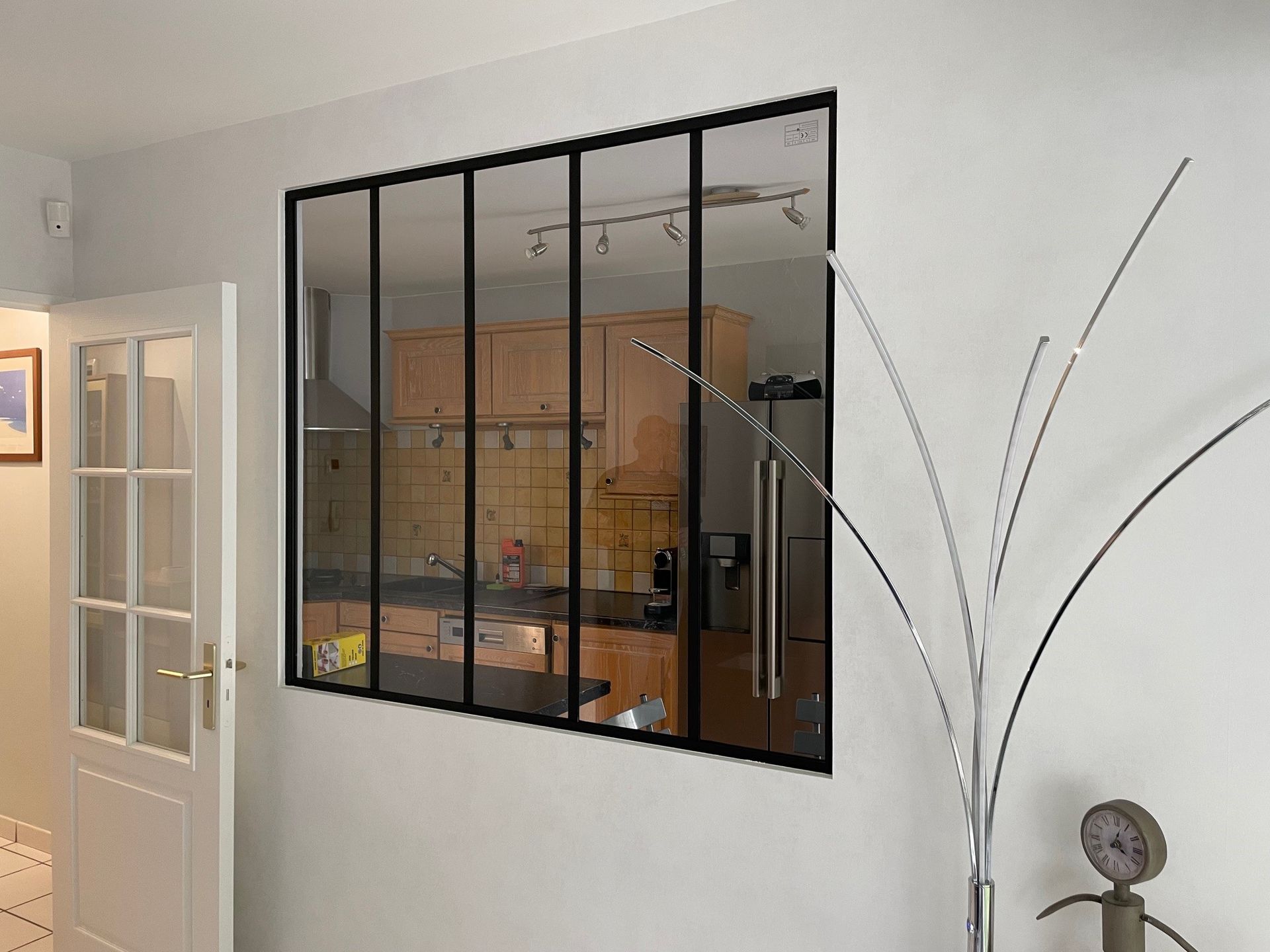 Fenêtre à cadre noir donnant sur une cuisine équipée de placards, d'une cuisinière et d'un réfrigérateur. Murs blancs.