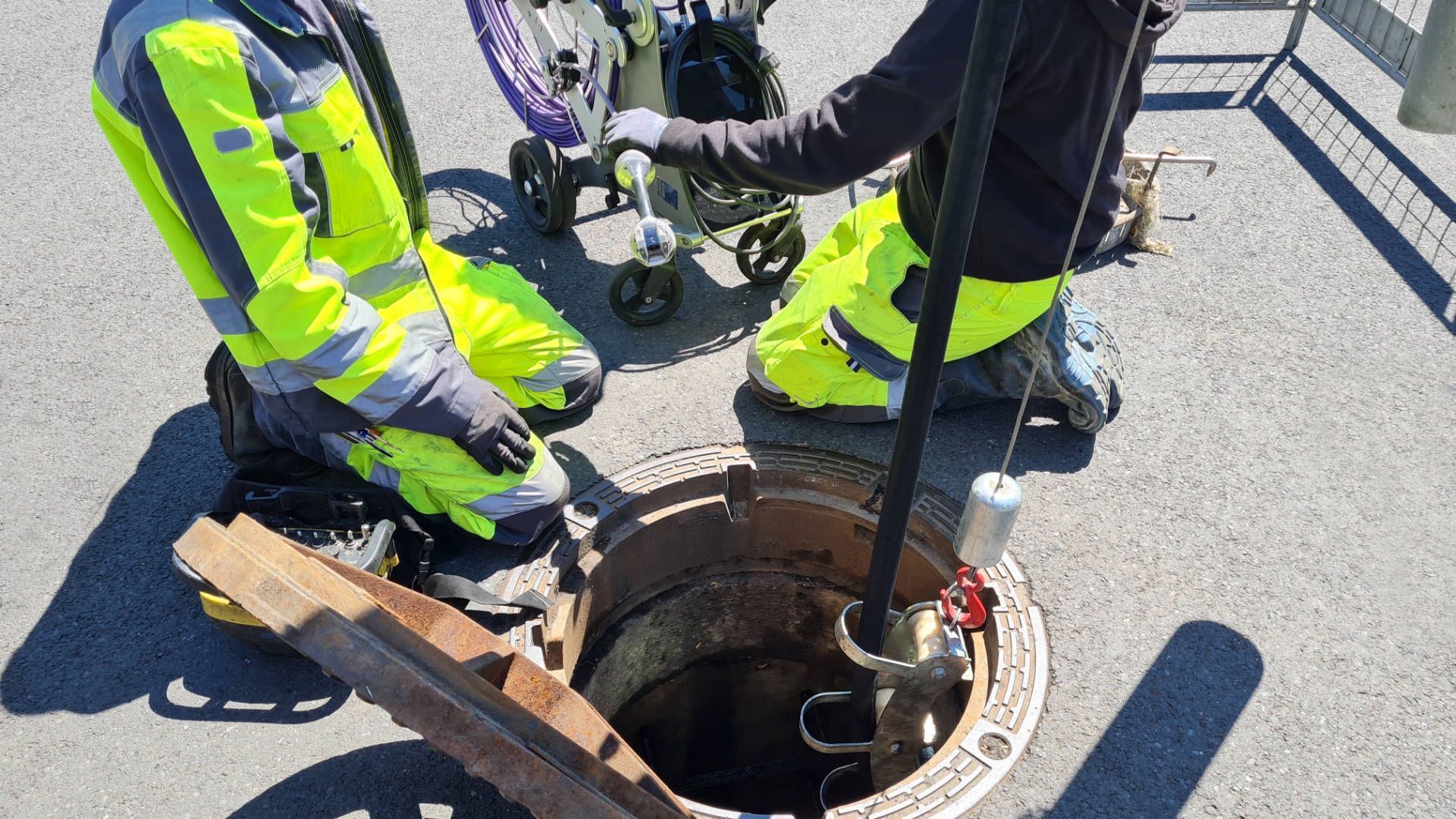 Zwei Arbeiter in Warnwesten untersuchen mit Werkzeugen einen Schachtdeckel. Im Freien auf dem Gehweg.