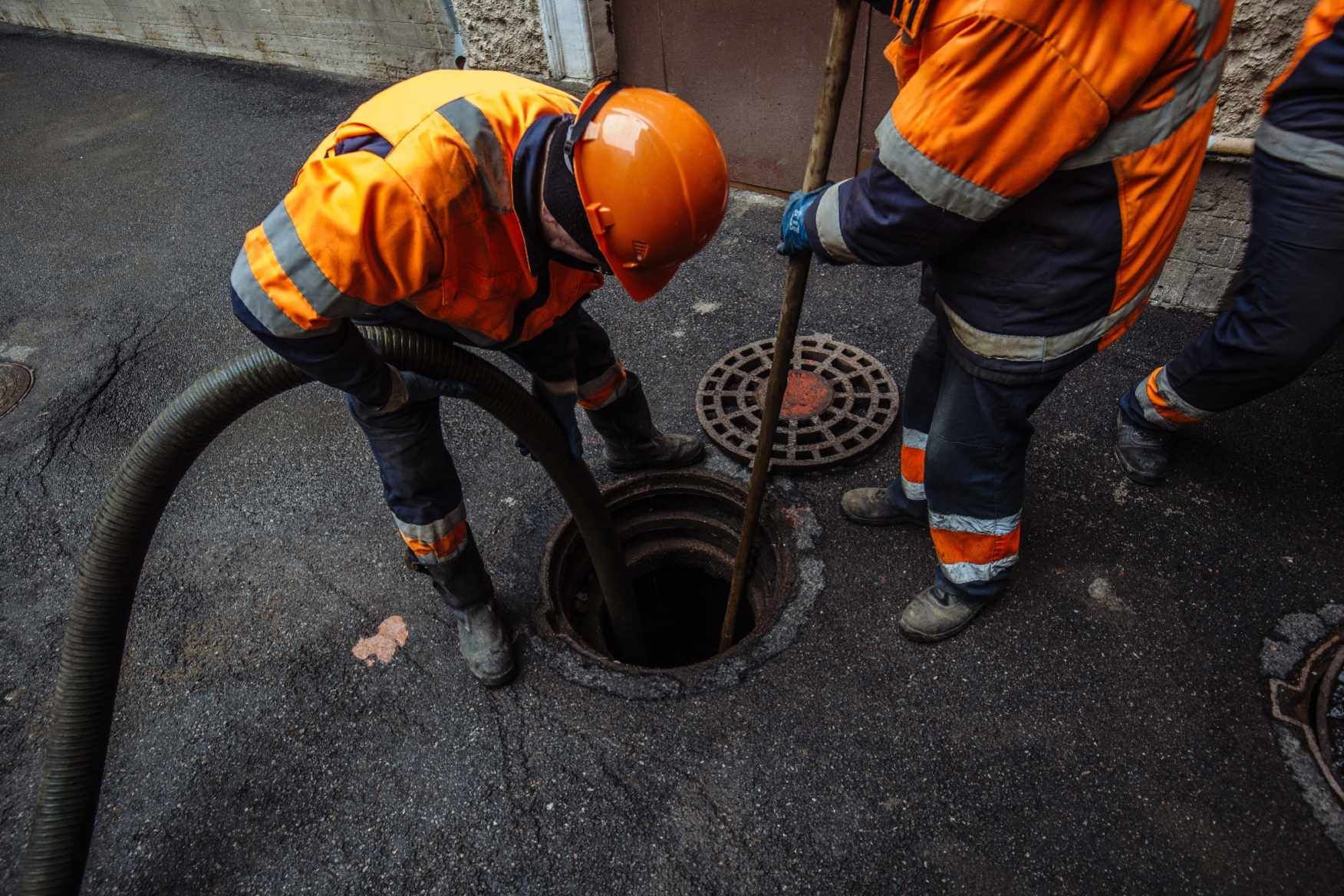 Arbeiter in orangefarbener Schutzkleidung reinigen einen Abwasserkanal in einer dunklen Straße.
