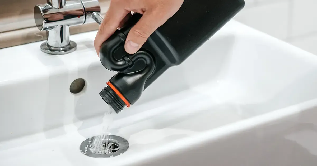 Eine Person gießt Wasser aus einem schwarzen Behälter in ein weißes Waschbecken.