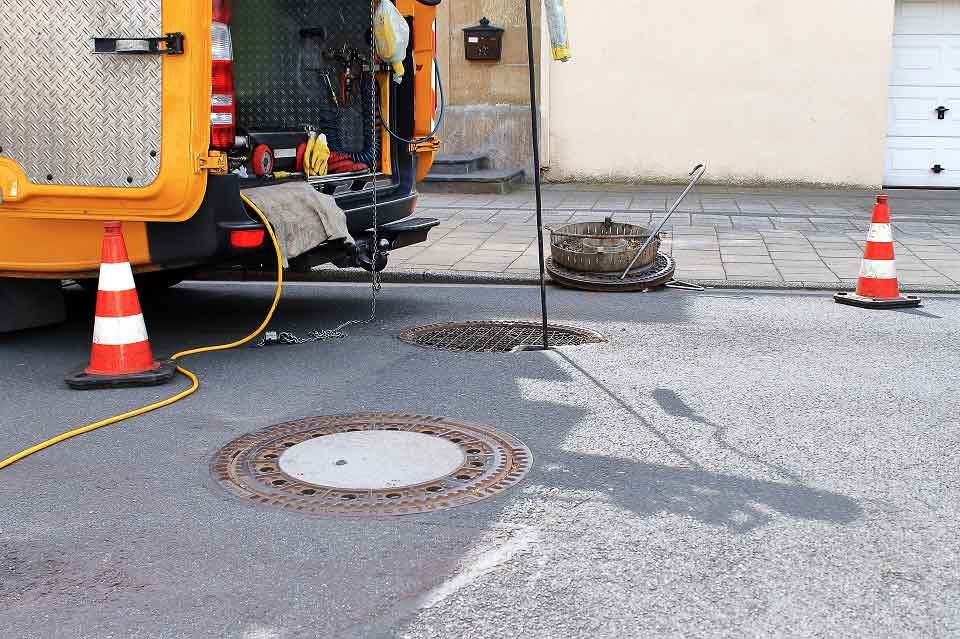 Kanaldeckel entfernt; Lieferwagen mit Leitkegeln auf der Straße geparkt; Werkzeuge sichtbar.