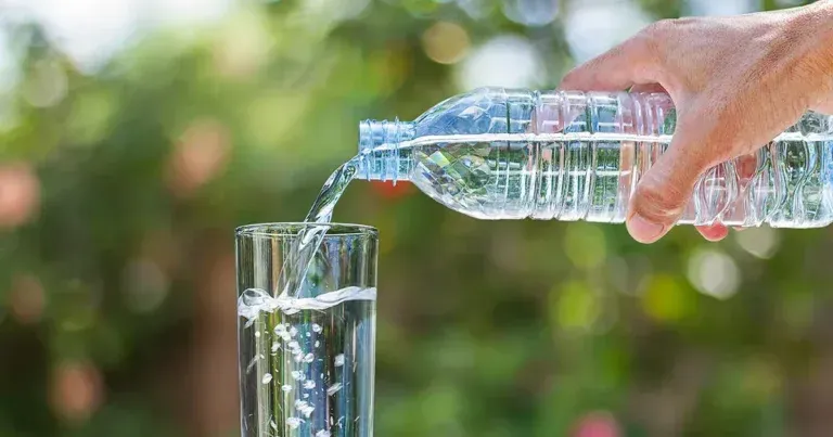 Eine Hand gießt Wasser aus einer Plastikflasche in ein klares Glas, im Freien, grüner Hintergrund.