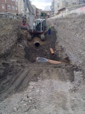 Excavadora cavando zanja para tuberías en un sitio de construcción, con edificios al fondo.