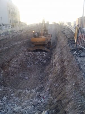 Excavadora amarilla en una zanja profunda en un sitio de construcción; edificios residenciales en el fondo.
