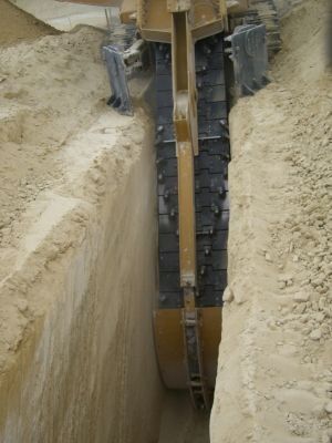 Una zanjadora cava una zanja estrecha en tierra de color claro; hay una persona cerca.