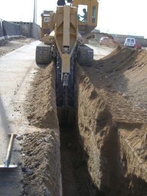 Maquina zanjadora cavando una zanja estrecha en un camino de tierra, con el operador visible en la cabina.