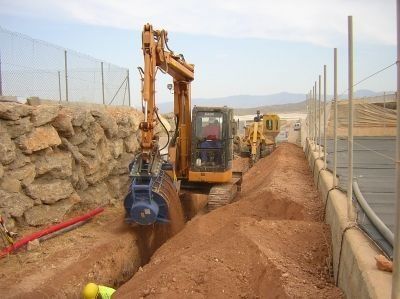 Excavadora cavando una zanja junto a un muro; obra. Tierra amarilla y marrón, cielo.