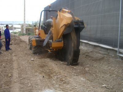 Zanjadora amarilla cortando tierra cerca de un edificio, trabajador conduciéndola, hombres observando.