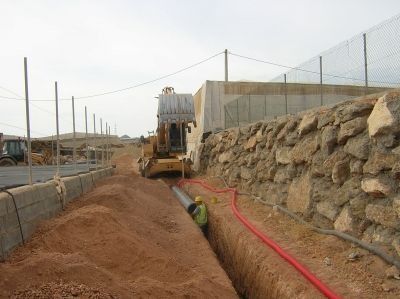 Sitio de construcción: excavadora, trabajador en zanja colocando tuberías, al lado de un muro de roca y una cerca de alambre.