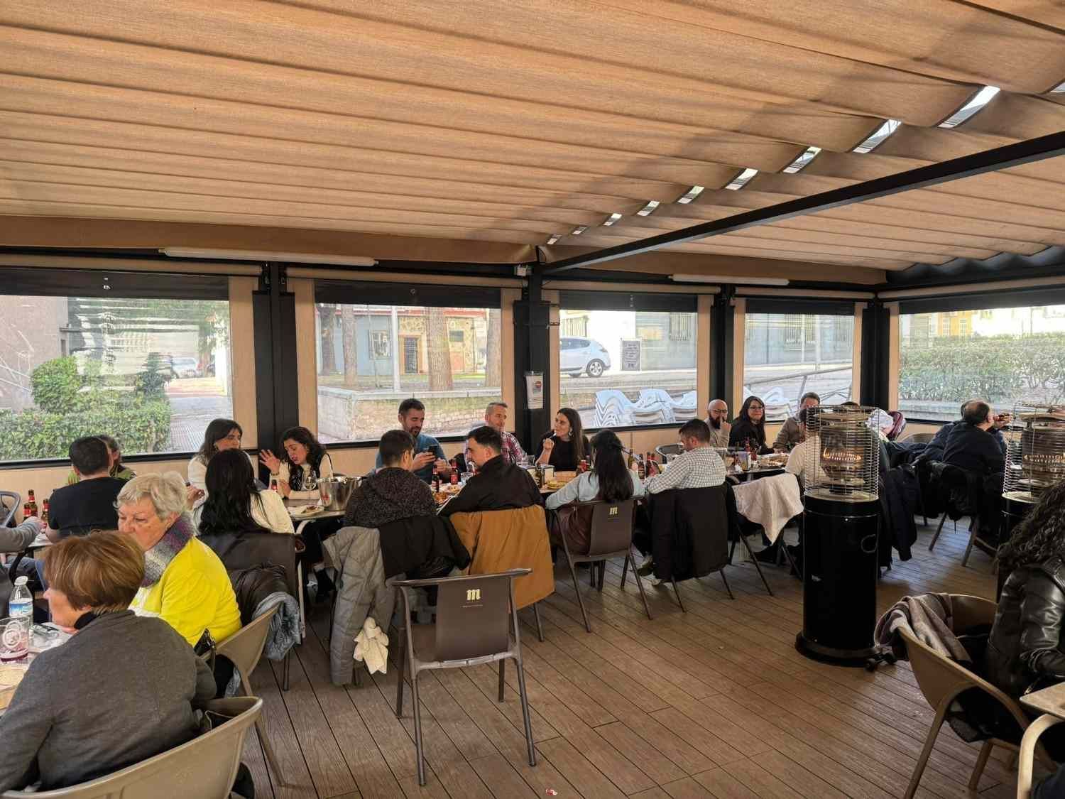 Personas sentadas a las mesas de una cafetería al aire libre con techo, vigas de madera y ventanales.