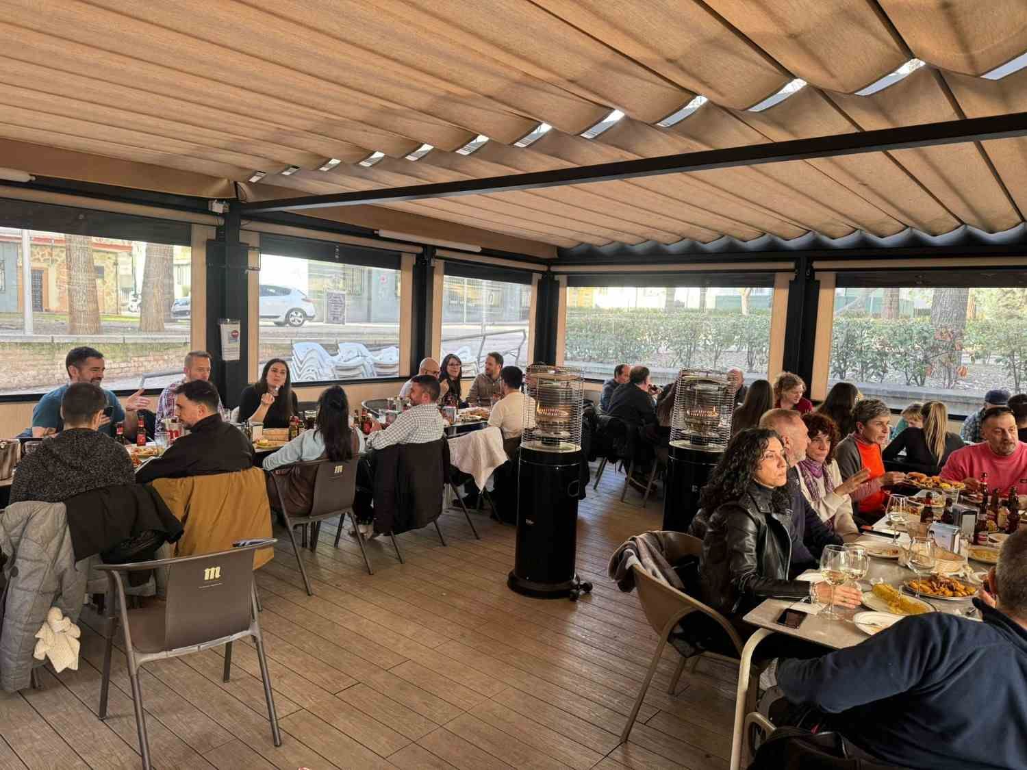 Personas comiendo en mesas largas en un restaurante al aire libre cubierto, con vigas de madera y grandes ventanales.