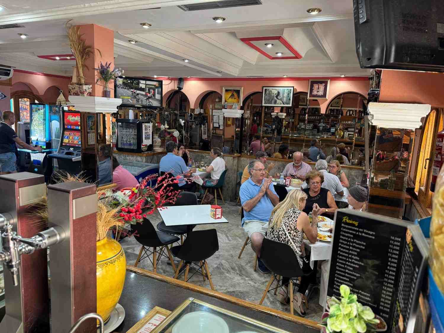 Cafetería o restaurante concurrido, con clientes sentados, máquinas recreativas y detalles decorativos en tonos rosa