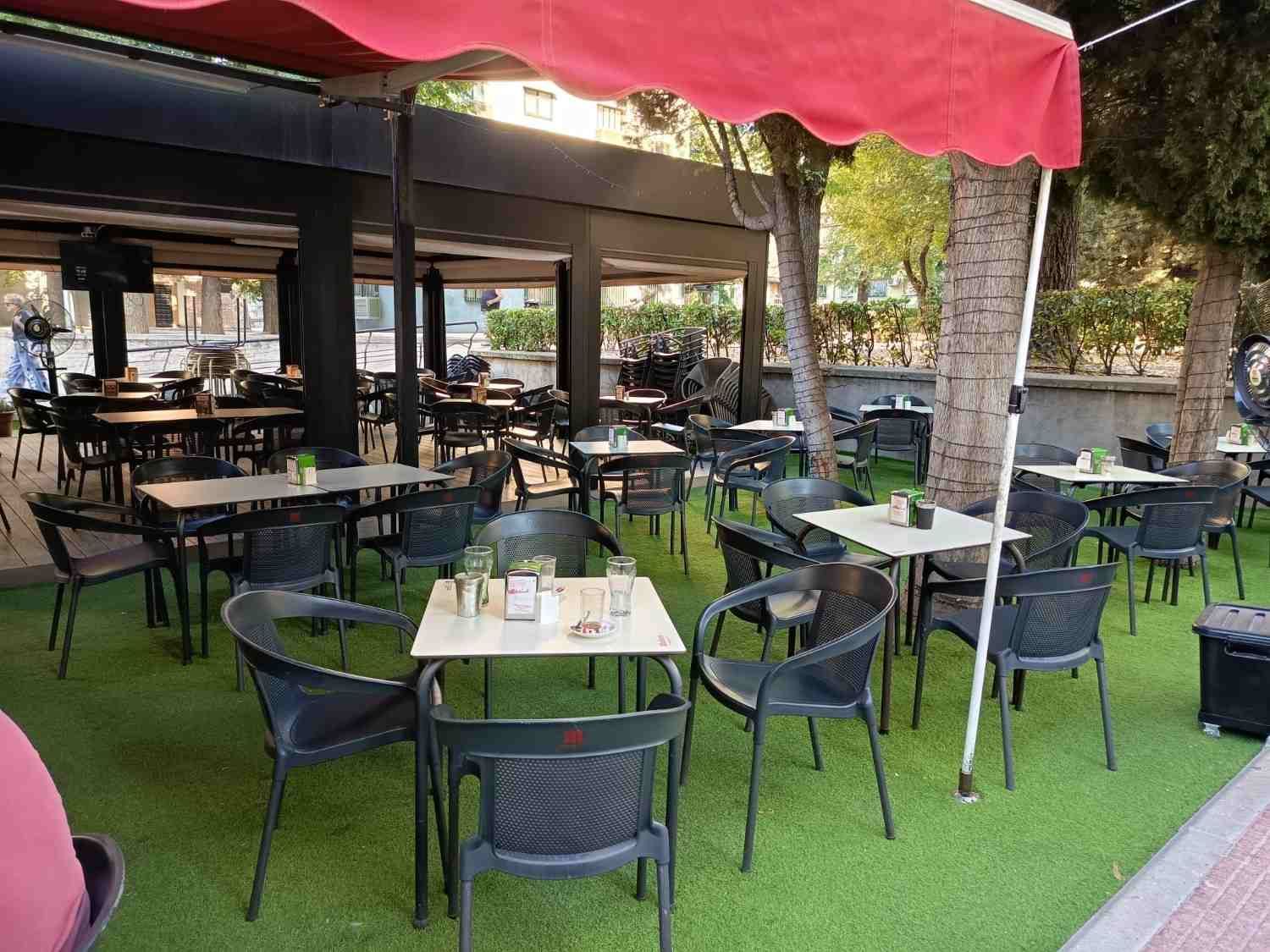 Terraza de cafetería al aire libre con sillas negras, mesas blancas y un toldo rojo sobre césped artificial