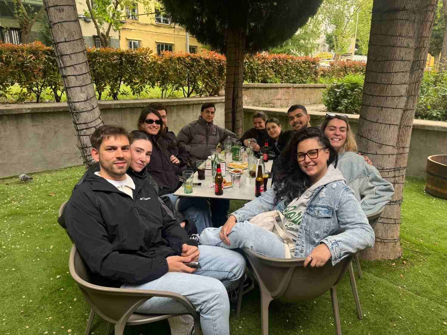 Un grupo de personas sentadas alrededor de una mesa al aire libre en un jardín, sonriendo y charlando bajo los árboles.