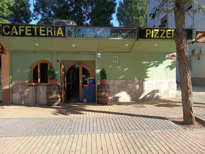 Fachada de cafetería y pizzería, de color verde y con una entrada arqueada, bajo los árboles