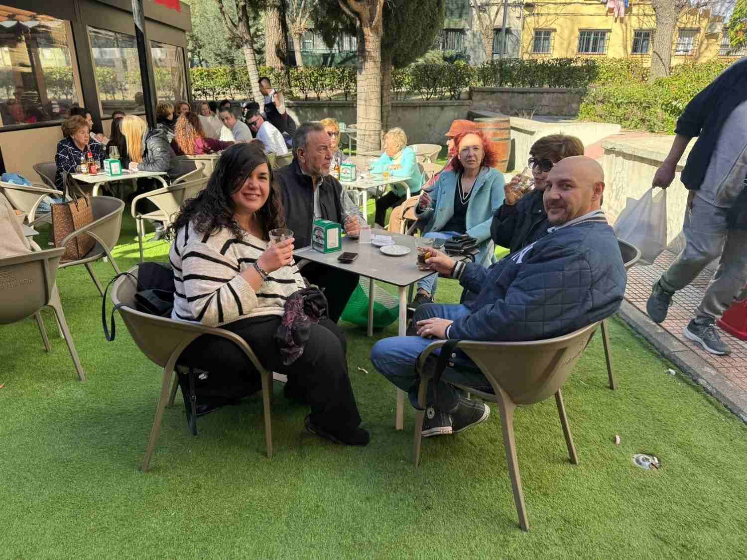 Personas sentadas en las mesas de una cafetería al aire libre en un jardín soleado, charlando y tomando café.