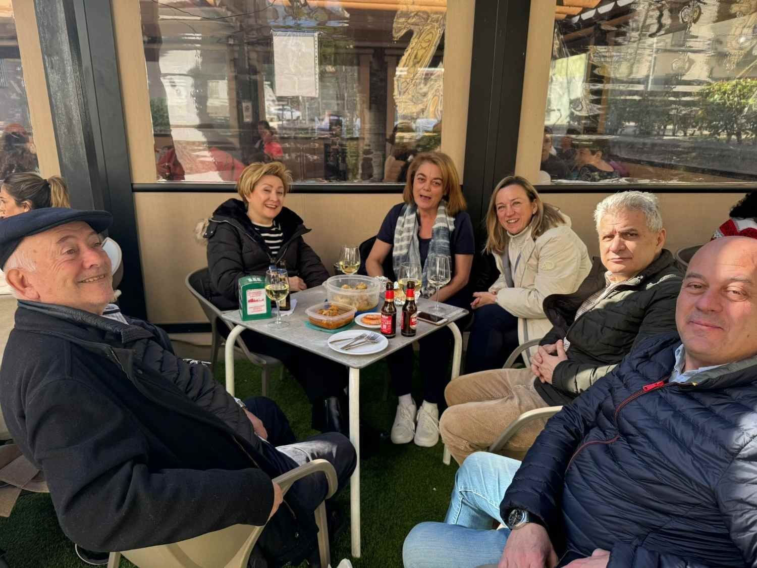 Seis personas sentadas en la mesa de una cafetería al aire libre, con bebidas y aperitivos, sonriendo y charlando