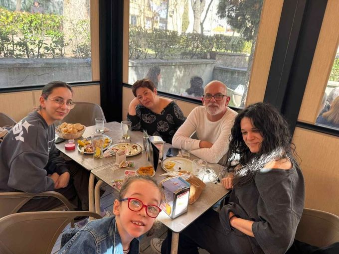 Cinco personas están sentadas alrededor de una mesa de cafetería con comida y bebidas, sonriendo junto a unos ventanales.