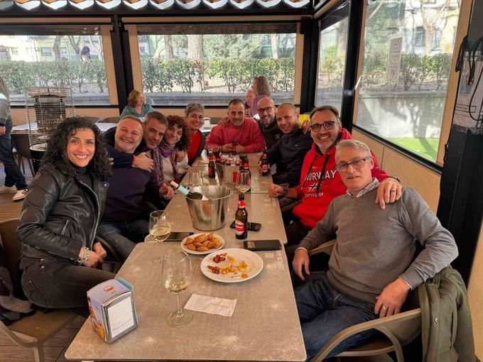 Grupo de personas sonriendo alrededor de una mesa larga en una cafetería acristalada, con platos, bebidas y una jarra metálica.