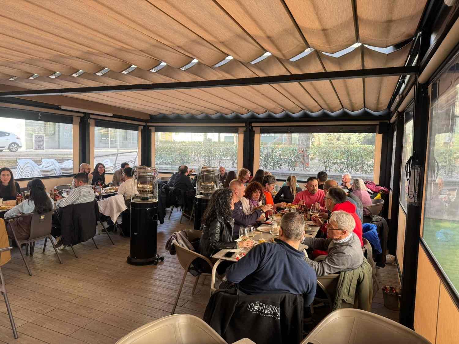 Personas sentadas a las mesas de un restaurante al aire libre cubierto, con barandillas negras y un toldo beige, comiendo juntas