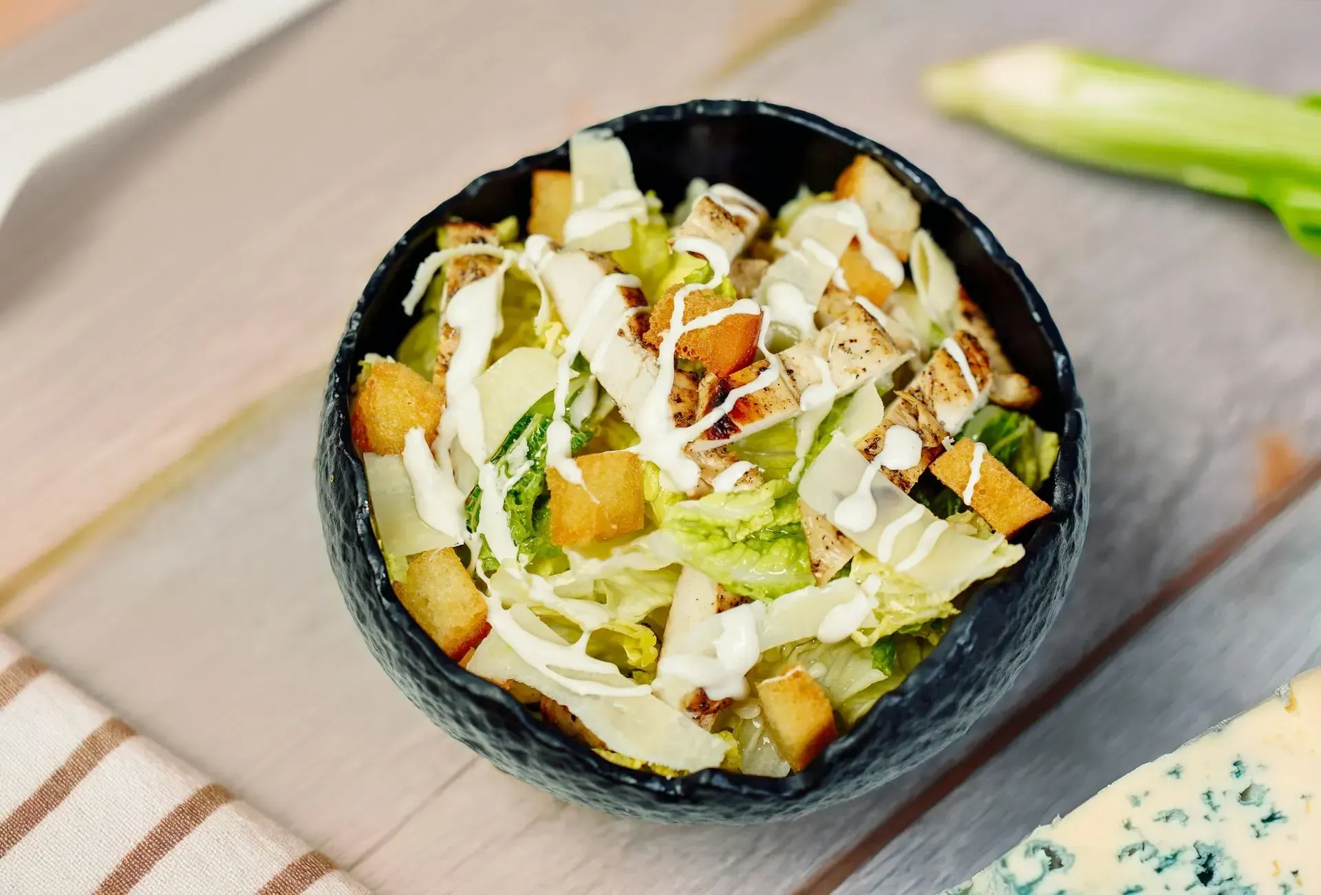 Una ensalada César con pollo a la parrilla, picatostes y virutas de parmesano en un bol negro sobre una mesa de madera.