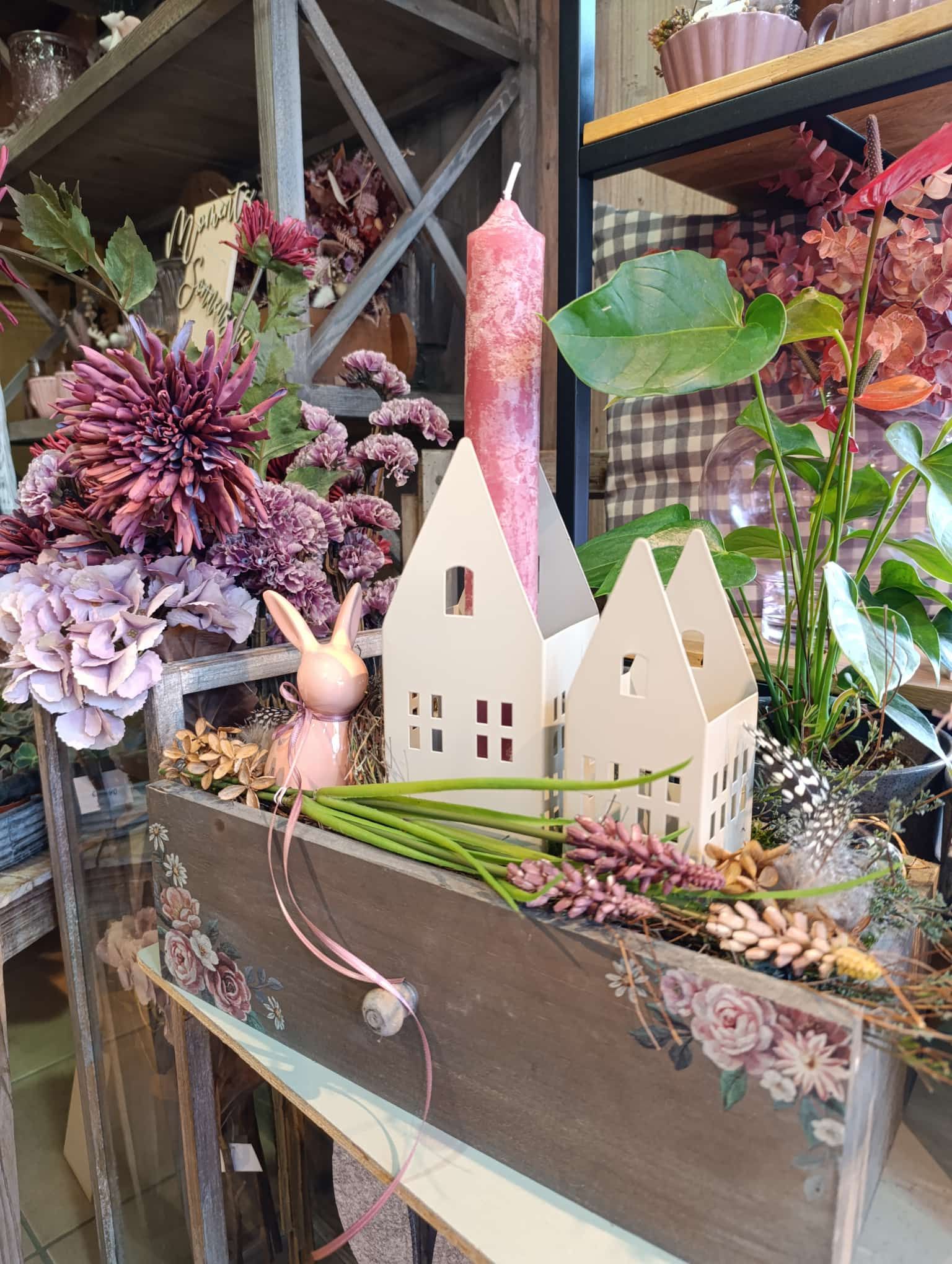 Eine Holzkiste gefüllt mit Blumen und einer Kerze in Form eines Hauses.