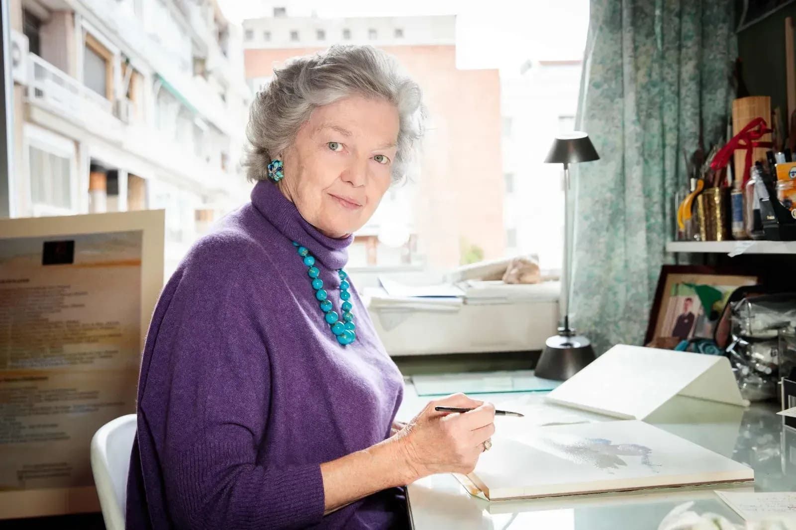 Una persona mayor, con el pelo gris, que lleva un jersey morado y un collar azul, está sentada en un escritorio dibujando en un cuaderno de bocetos.