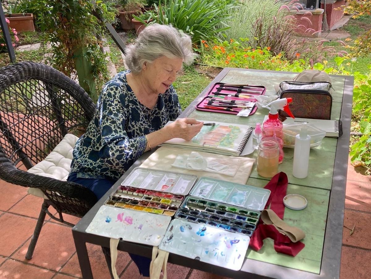 Una anciana está sentada en una mesa pintando con acuarelas.