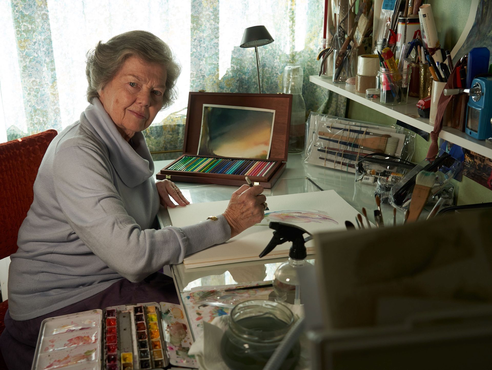 Una anciana está sentada en un escritorio pintando con acuarelas.