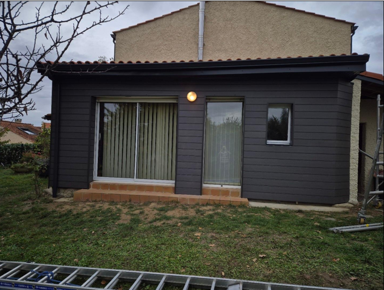 Petite maison gris foncé avec de grandes fenêtres et une lampe extérieure allumée dans une cour gazonnée.