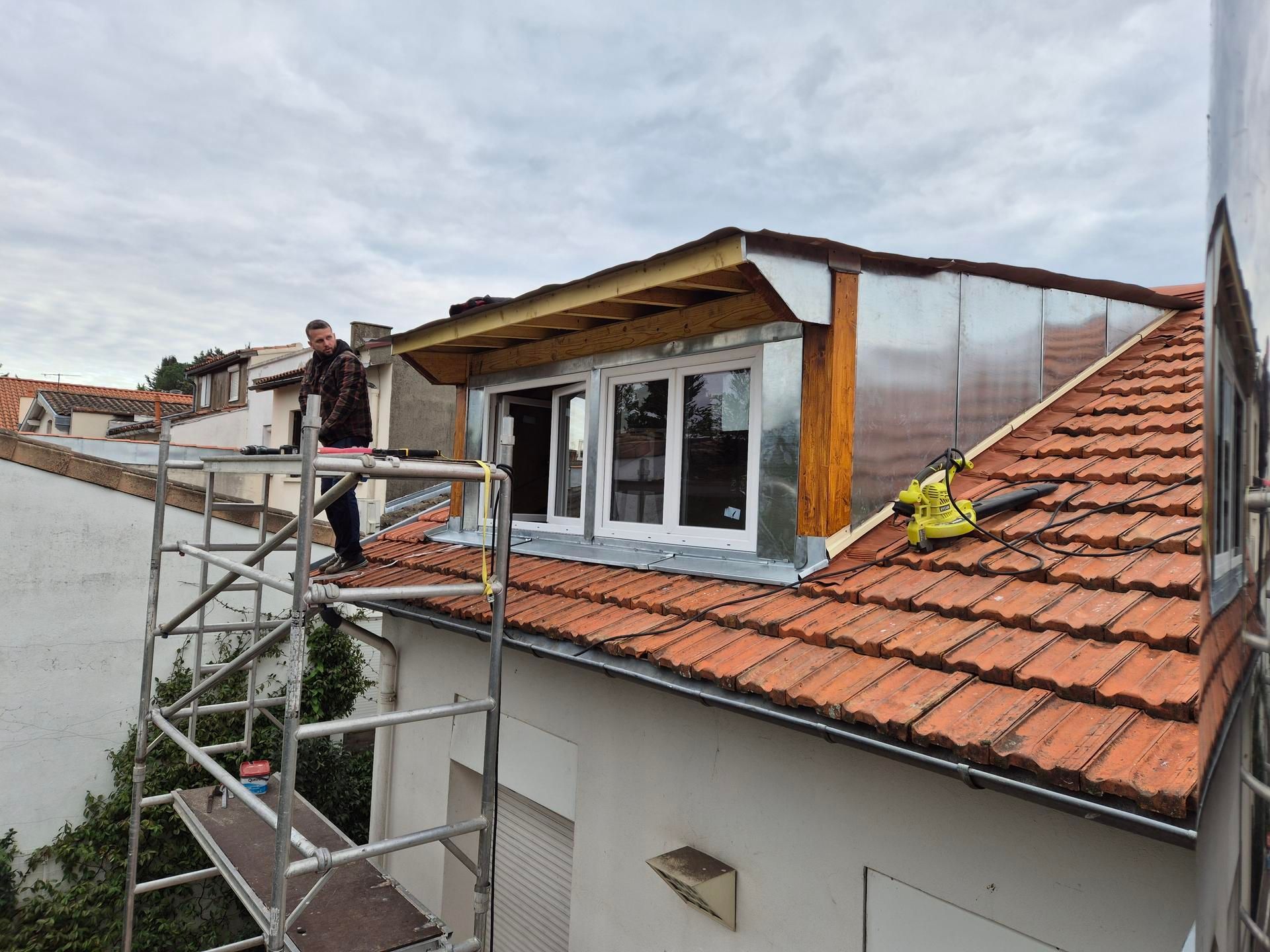 Un ouvrier sur un échafaudage répare le toit d'une lucarne avec des tuiles rouges et des fenêtres blanches sur une maison.