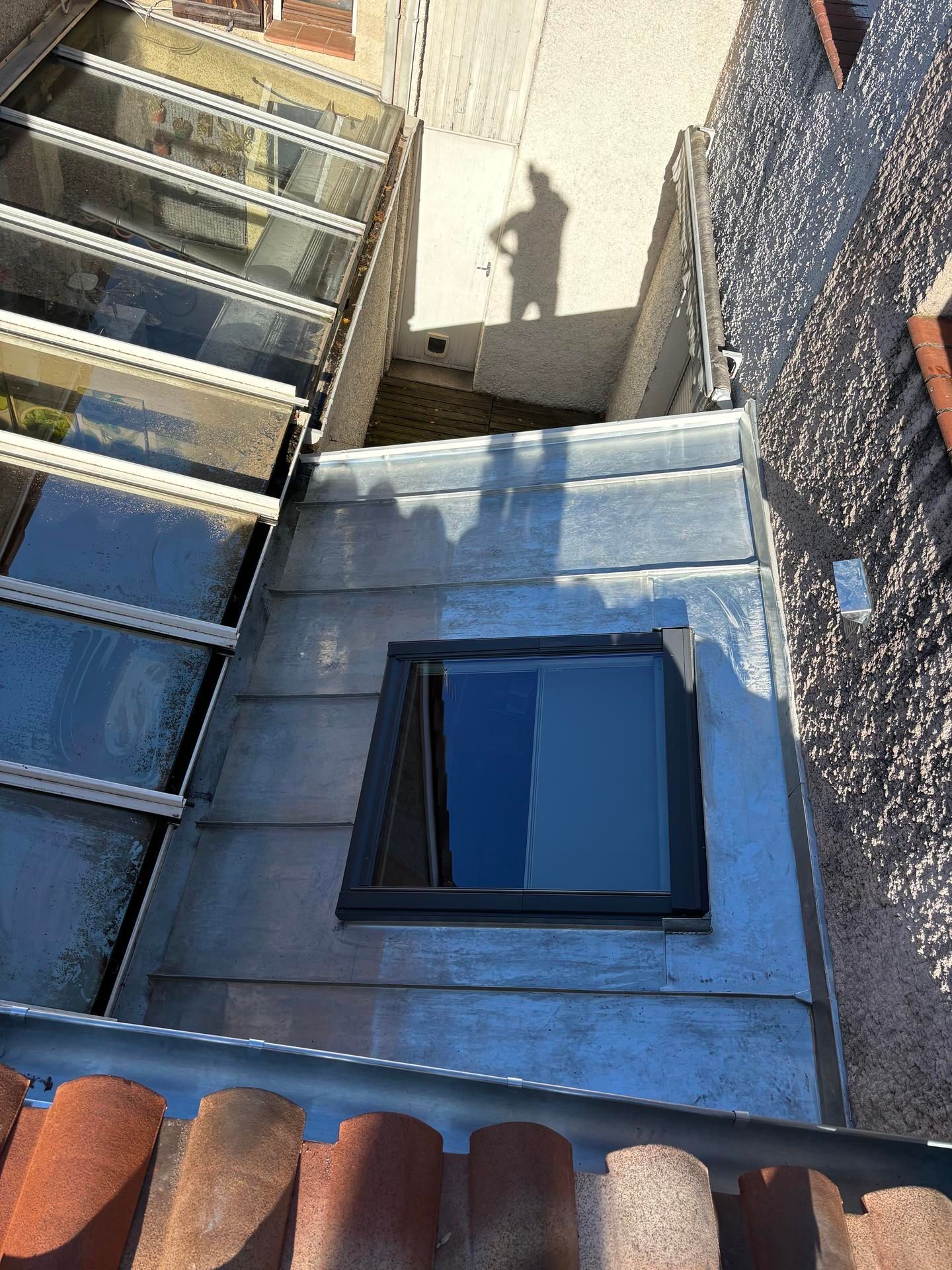 Escalier menant au toit, avec fenêtre bleue, toit gris et l'ombre d'une personne projetée sur le mur.
