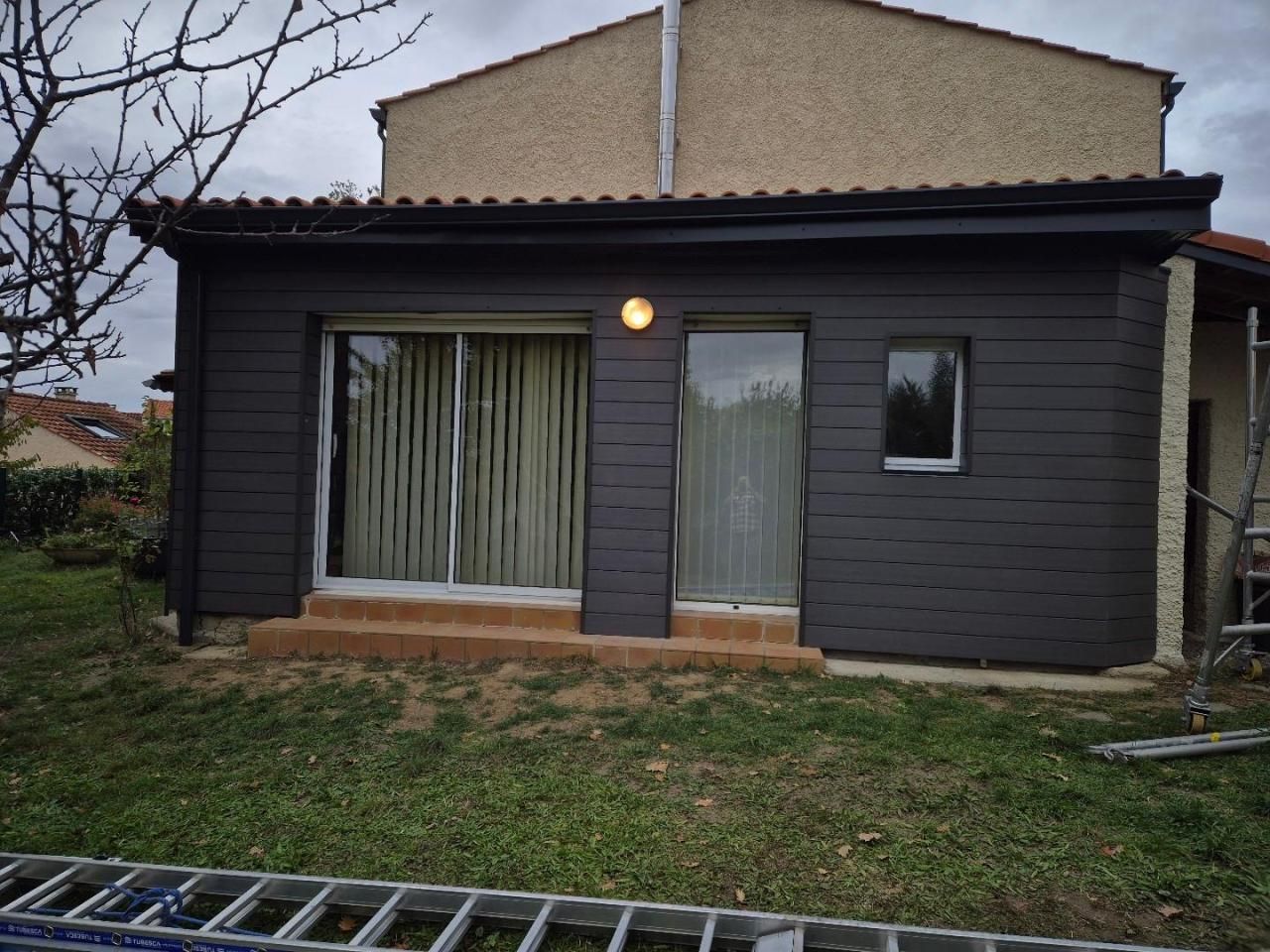 Extérieur d'une petite maison sombre avec de grandes fenêtres, une porte d'entrée et une cour herbeuse sous un ciel couvert.