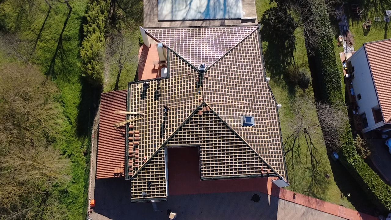 Vue aérienne d'une maison au toit partiellement recouvert de tuiles, entourée d'arbres et d'une allée.