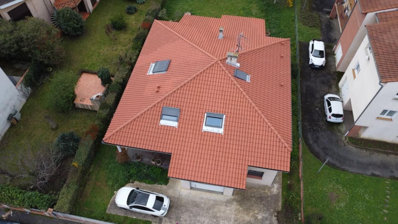 Vue aérienne d'une maison aux tuiles rouges avec plusieurs fenêtres de toit et des voitures garées à côté.