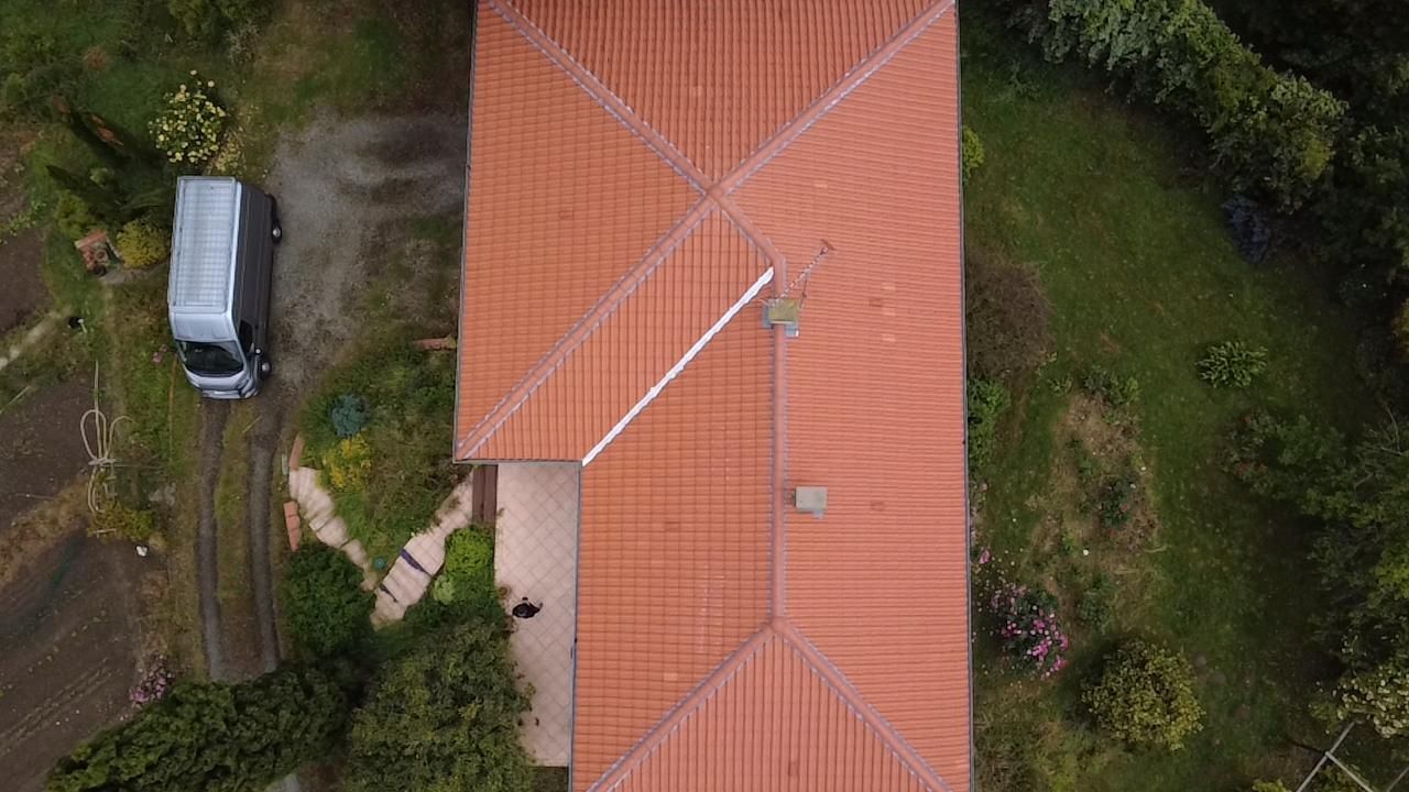Vue aérienne d'une maison au toit de tuiles rouges à côté d'une camionnette garée sur un chemin de terre, entourée de verdure.