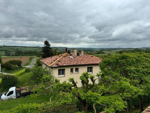 Maison avec un toit en tuiles rouges sous un ciel nuageux, entourée de champs verdoyants et d'arbres ; camionnette stationnée dans l'allée.
