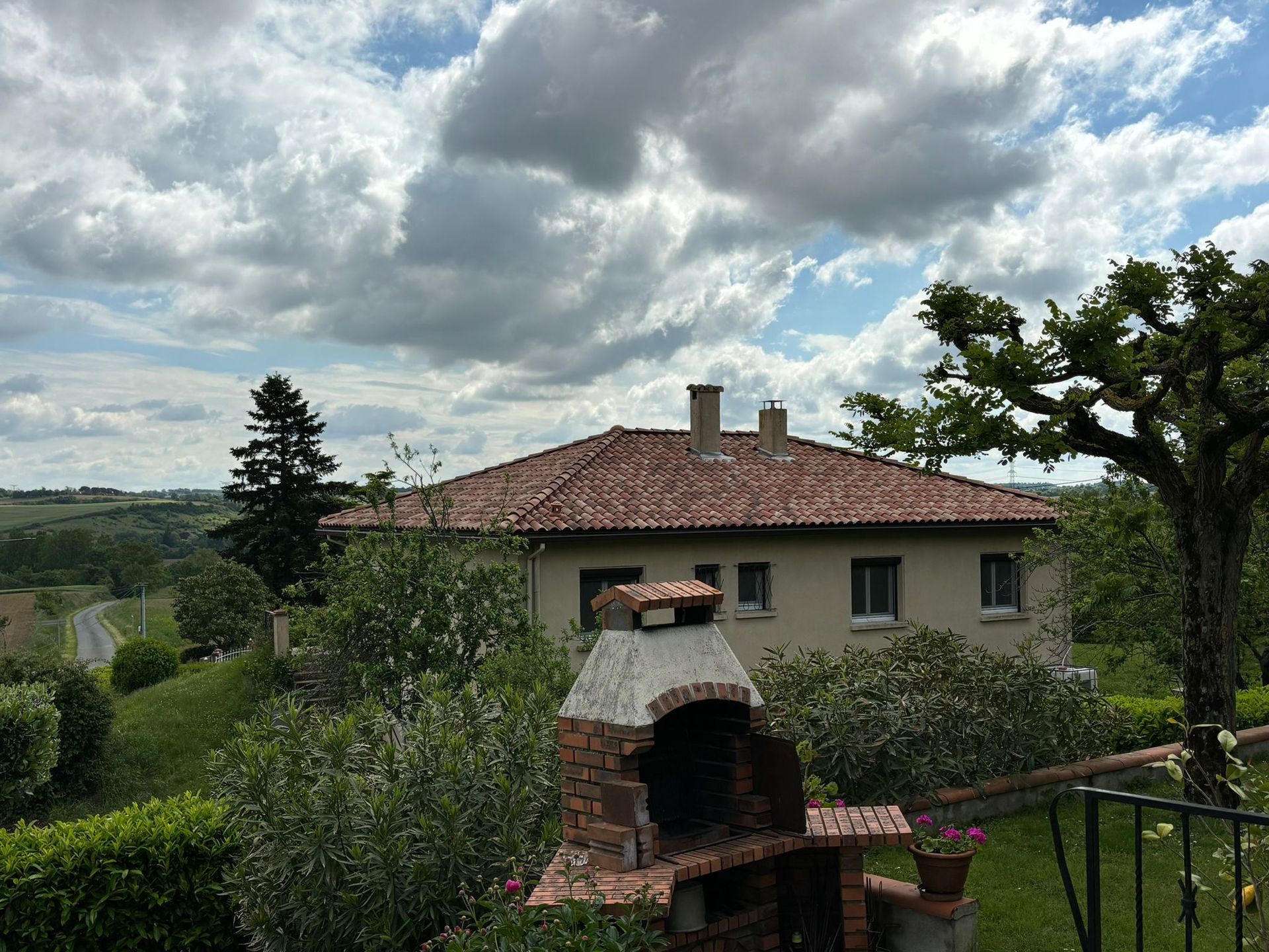 Maison avec un toit en tuiles, barbecue en briques au premier plan, et ciel nuageux au-dessus de collines verdoyantes.