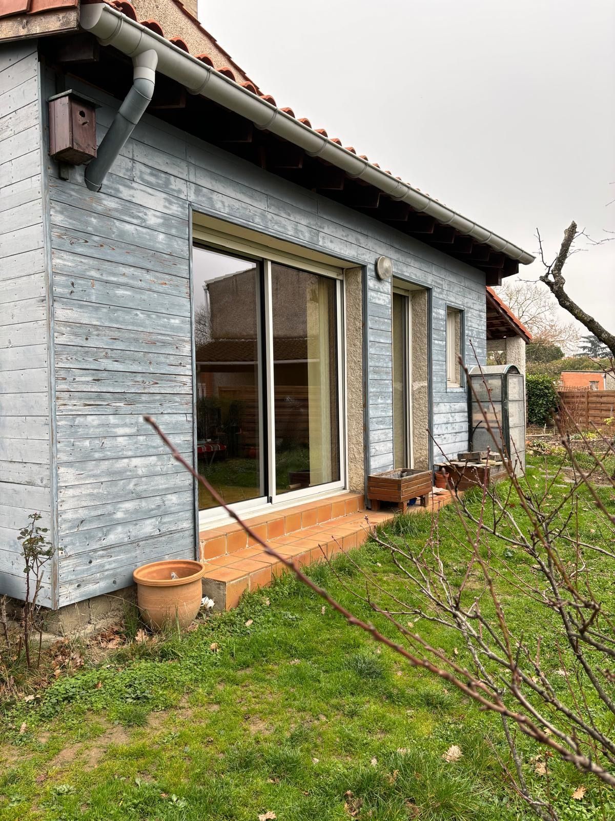 Maison à la façade grise avec portes coulissantes en verre, petite terrasse et jardin gazonné.