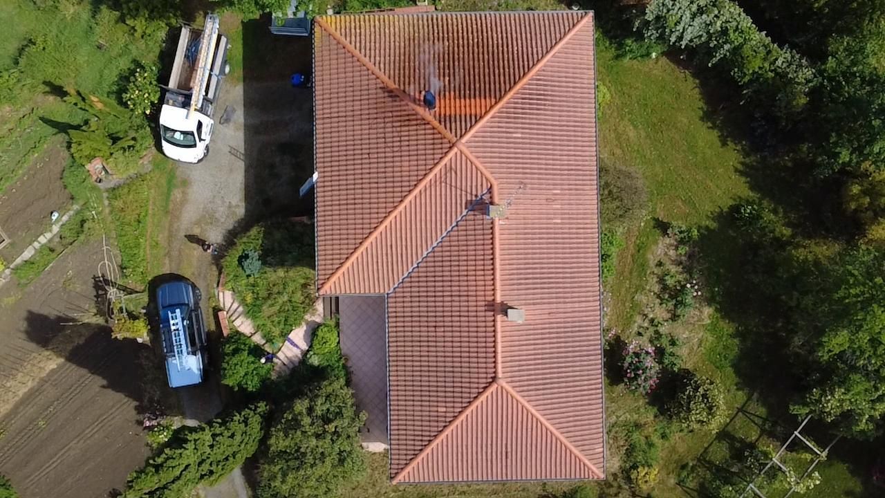 Vue aérienne d'une maison au toit de tuiles rouges, entourée d'herbe, d'arbres et de deux voitures garées.