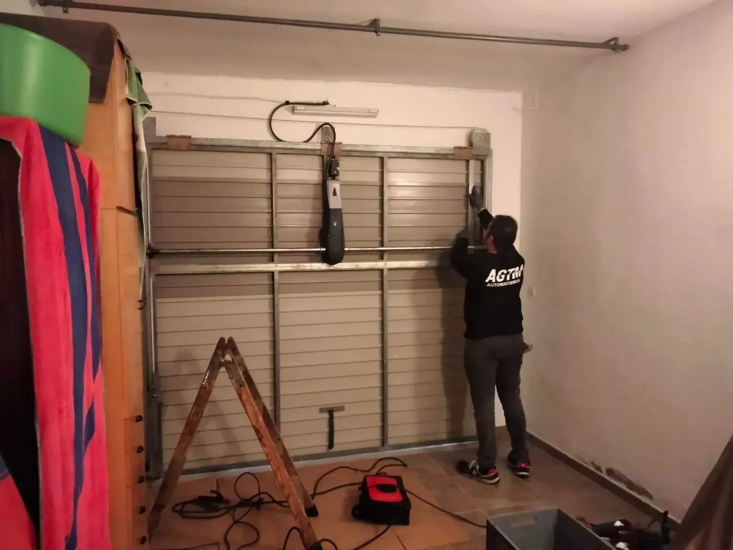 Una persona reparando una puerta de garaje. Está en el garaje, de pie en el suelo, usando una herramienta.