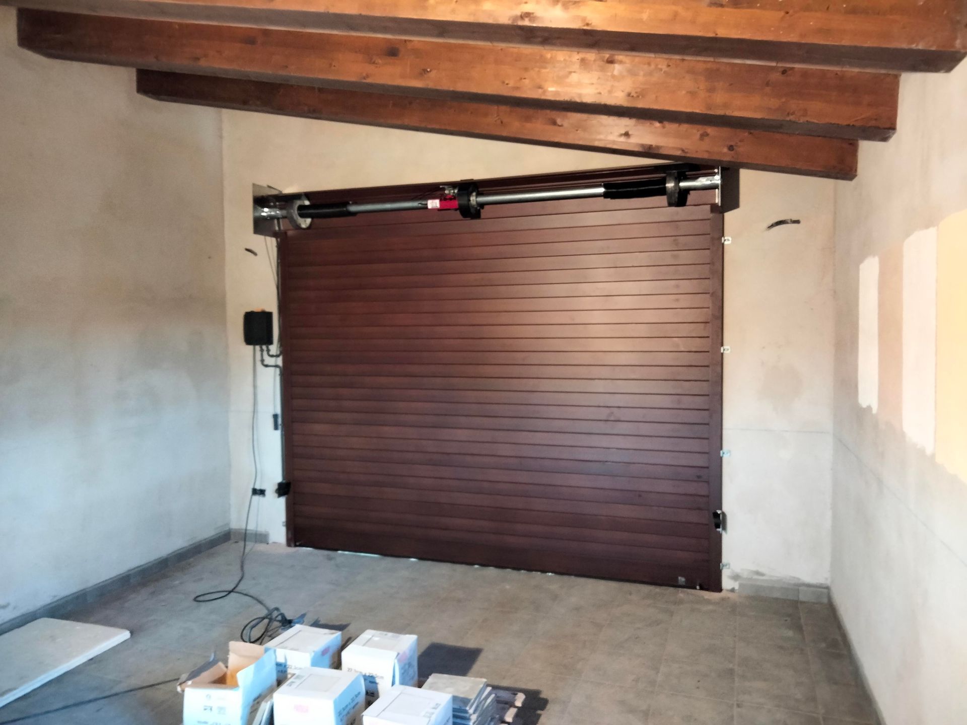 Puerta de garaje marrón de lamas horizontales, parcialmente abierta. Vigas de madera, techo visto y suelo de hormigón.