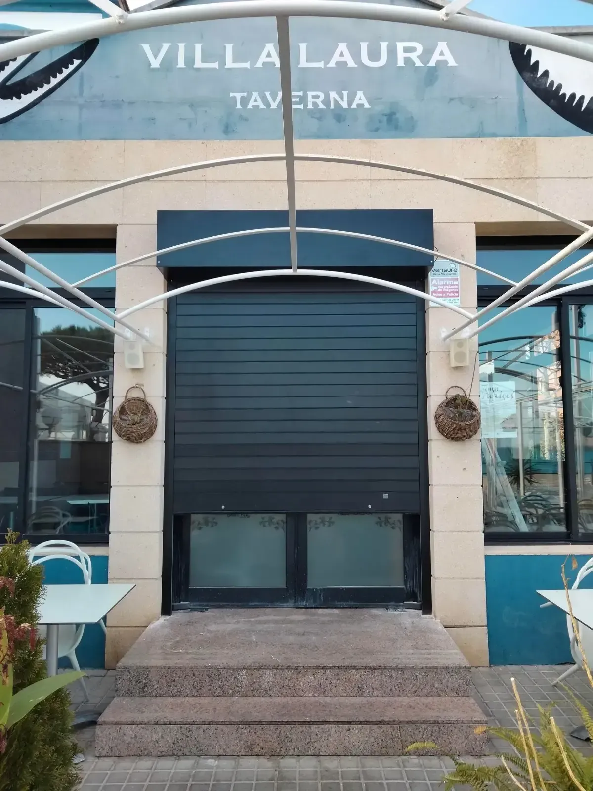Entrada de la taberna Villa Laura con puerta negra cerrada, fachada color crema, ventanas de vidrio y detalles azules.