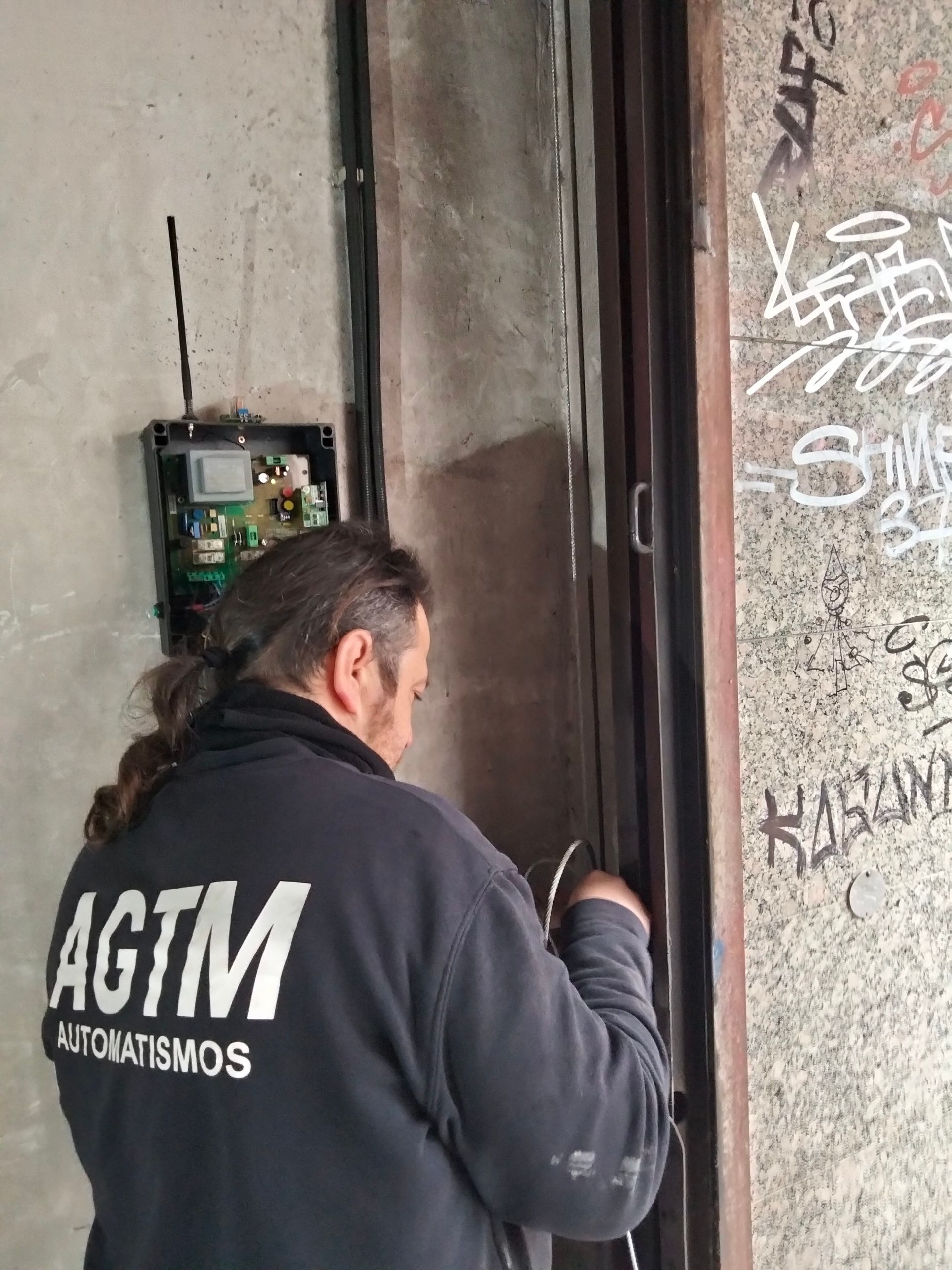 Una persona con una sudadera negra AGTM trabaja en una caja de control cerca del marco de una puerta, cerca de un grafiti.