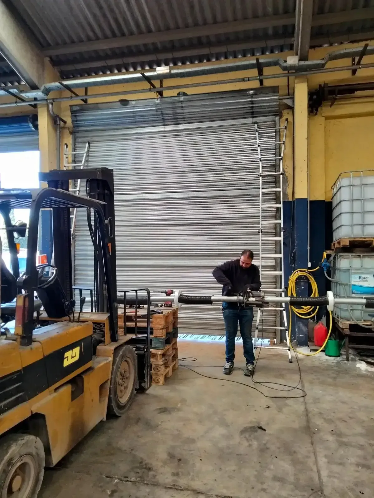 Hombre trabajando con maquinaria cerca de una puerta de garaje cerrada y una carretilla elevadora dentro de un almacén.