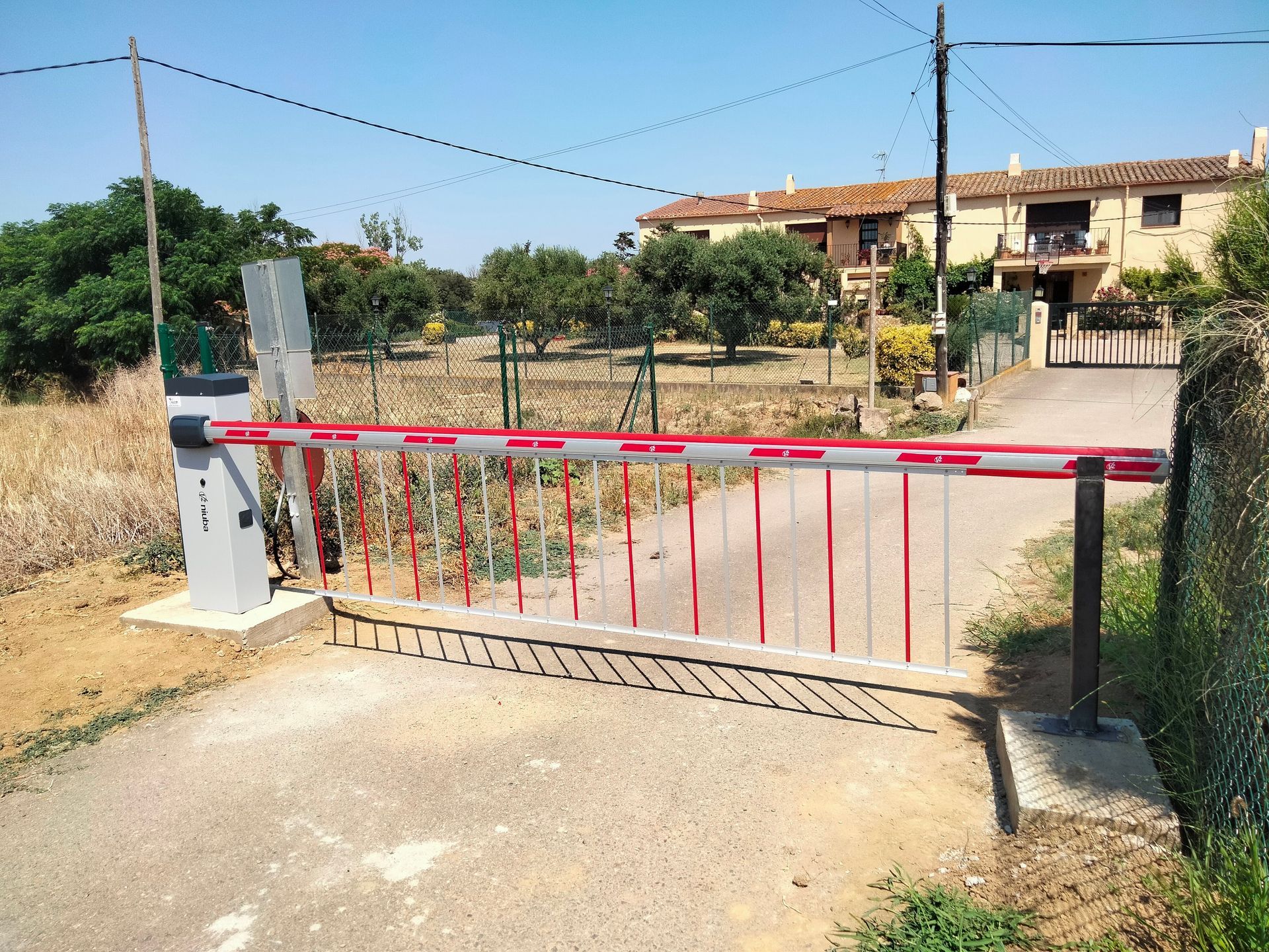 Barrera de rayas rojas y blancas que bloquea un camino rural, con edificios al fondo.