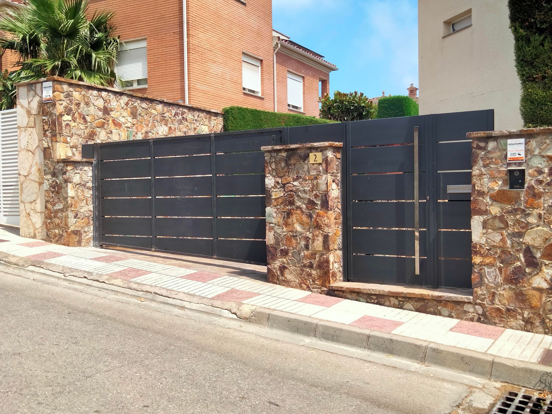 Portón de piedra y metal en entrada residencial; portones de metal gris, muros de piedra y acera de ladrillo.