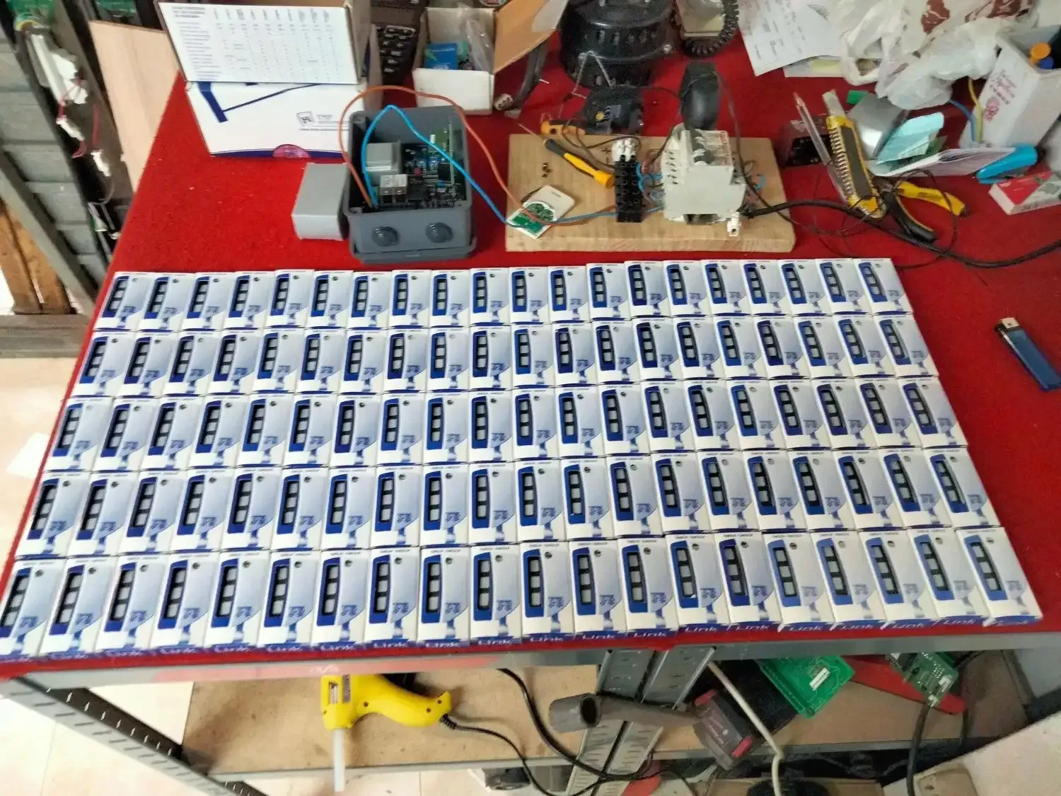 Una mesa roja con muchos elementos idénticos en cajas azules y blancas, posiblemente dispositivos electrónicos y algunas herramientas.