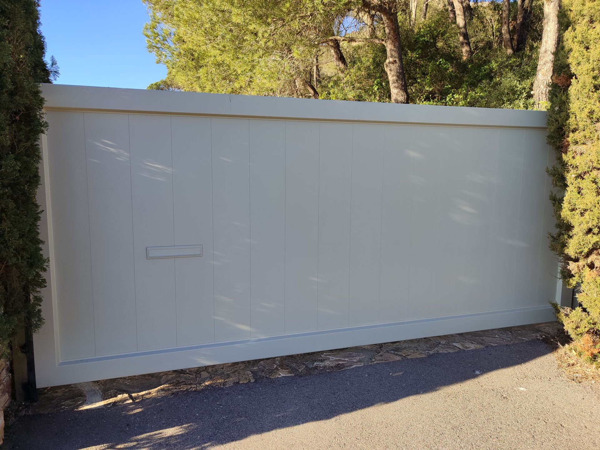 Puerta de madera gris situada en una entrada, flanqueada por setos verdes, bajo un cielo soleado.